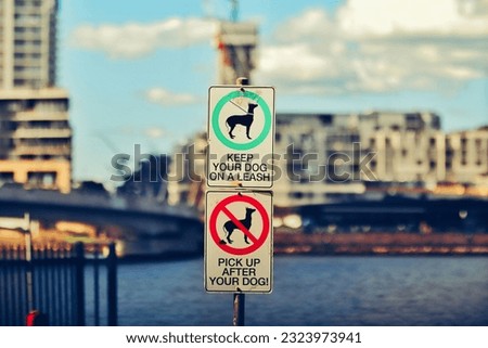 Two street signs of "keep your dog on a leash" and "pick up after you dog" with a cityscape on the background