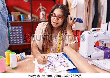 Young beautiful arab woman tailor listening voice message by smartphone writing on notebook at atelier
