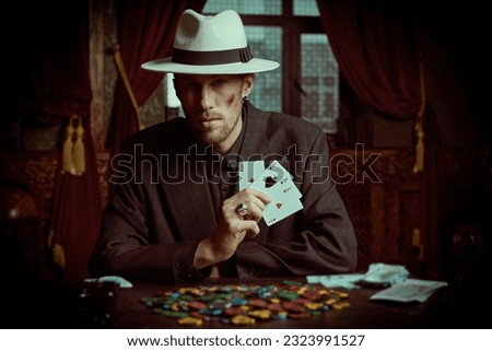 The criminal world of the mafia. A tough gangster man in an elegant black suit and a white hat sits in a luxury apartment and plays cards. Retro style. Detective movie. Royalty-Free Stock Photo #2323991527