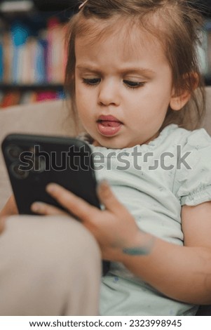 Cute little girl in casual clothes smiling and looking at camera while sitting on sofa and using smartphone