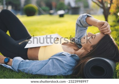 Side view of young woman lying on grass and reading book while resting in park on sunny day