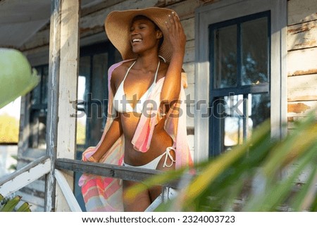 Happy african american woman in bikini and sunhat standing on porch of beach house, copy space. Summer, fashion and beauty, free time and vacations, unaltered.