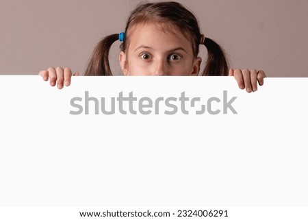 Happy pretty girl smiling and posing hiding behind empty whiteboard. Child holding paper poster advertising Your text. 