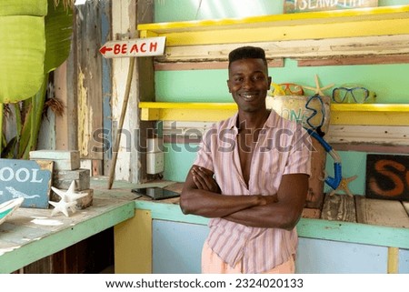 Portrait of happy african american man standing behind counter of surf hire beach shack, copy space. Local business, business owner, hobbies, sport, surfing, summer and vacation, unaltered.