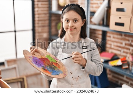 Young woman with down syndrome smiling confident holding paintbrush and palette at art studio Royalty-Free Stock Photo #2324024873