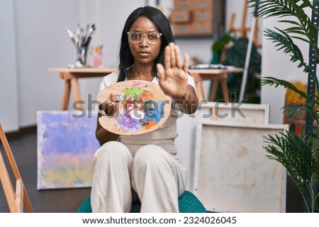 African young woman holding painter palette with open hand doing stop sign with serious and confident expression, defense gesture 
