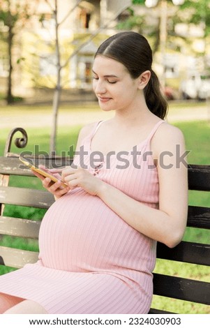pregnant woman sits on a park bench and texting on phone.