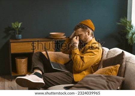 Sad and tired man is depicted sitting on a sofa at home. Overwhelmed by bad news or challenges, he holds his head with closed eyes in his hand, conveying a sense of emotional distress. High quality Royalty-Free Stock Photo #2324039965