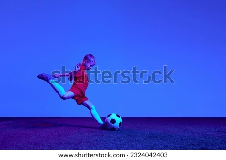 One sportive child, boy in red uniform playing, training football over dark blue background in neon. Concept of action, sportive lifestyle, team game, health, energy, vitality and ad