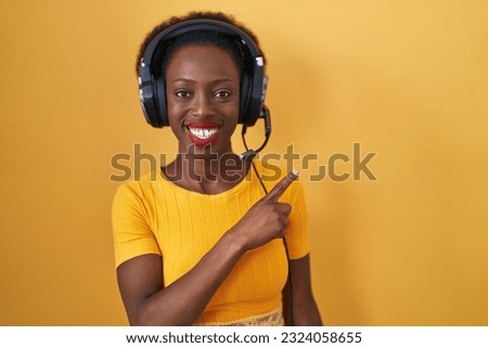 African woman with curly hair standing over yellow background wearing headphones cheerful with a smile of face pointing with hand and finger up to the side with happy and natural expression on face 