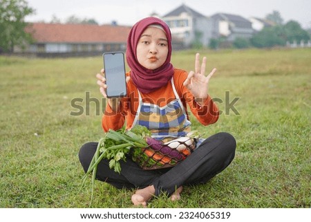 Asian woman in hijab wearing apron sitting carrying basket full of vegetables by holding blank white cell phone screen and gesturing okay, field background.