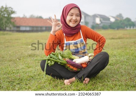 Smiling Asian woman in hijab wearing apron sitting holding basket full of vegetables and gesturing okay, field background.