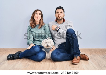 Young hispanic couple sitting on the floor with dog making fish face with lips, crazy and comical gesture. funny expression. 