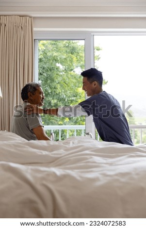 Happy diverse male doctor and senior male patient sitting on bed and discussing at home. Medical services, check up, home visit, healthcare, wellbeing, and senior lifestyle, unaltered.