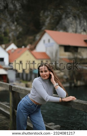 girl in spring or autumn near a beautiful view of the river and Gori, a small town on the background. the girl is dressed in a pink coat and blue jeans, smiling and happy