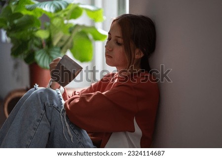 Frustrated sad teenage girl sitting on floor with cup devastated thinking about trouble, broken heart. Young woman have bad mood due to hormones in period. Depressive thoughts from problems at school. Royalty-Free Stock Photo #2324114687