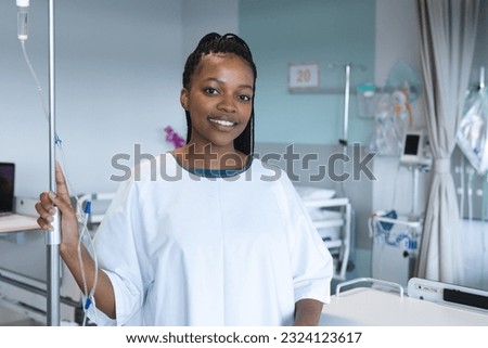Portrait of happy african american female patient with braids, wearing hospital gown, holding drip. Hospital, medicine, healthcare and work, unaltered. Royalty-Free Stock Photo #2324123617