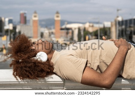 Relaxed young man lying in a balcony while listening music with headphones. Confident young guy lying down with closed eyes listening to the radio with the city landscape in the background.