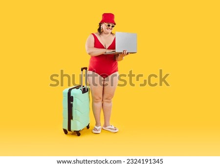 Happy funny fat woman in red hat and swimsuit with suitcase getting ready for holiday trip booking tickets online via laptop isolated on studio yellow background. Vacation and summer journey concept.