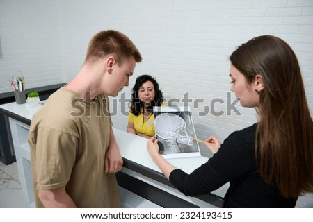 Young woman dentist doctor explaining X-ray image to a male patient, standing together at the reception counter of a modern medical clinic or outpatient hospital. Healthcare and medicine. Diagnostics Royalty-Free Stock Photo #2324193415