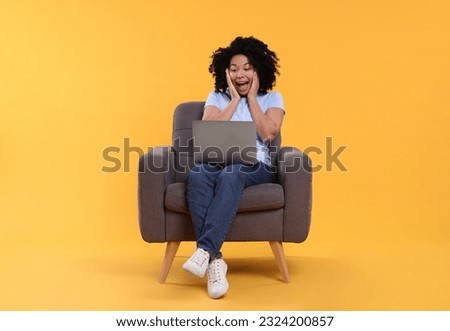 Emotional young woman with laptop sitting in armchair against yellow background