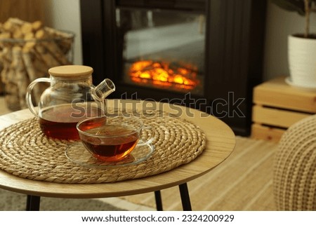 Teapot and cup of drink on coffee table near stylish fireplace in cosy living room. Interior design