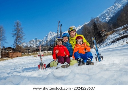 Family, dad son and little kids sit in the snow in sport outfit with skis helmets masks laugh hug smiling over mountain