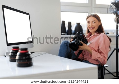 Happy lady photographer holding her DSLR camera, sitting at workplace with computer monitor with blank screen, working with post processing editing software on modern desktop
