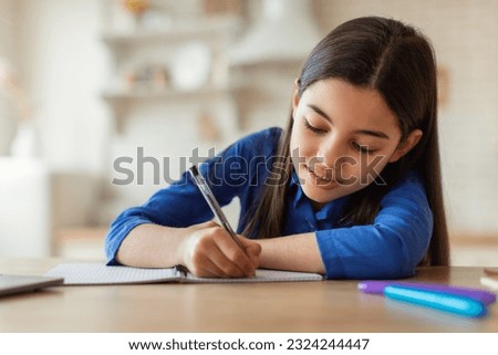 E-Learning. Arabic School Girl Writing Notes While Studying With Laptop Sitting At Table At Home. Cute Preteen Kid Doing Homework Online And Noting Information From Internet Lecture Near Computer