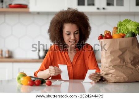 Market Delivery. Happy Black Lady Checking Bills In Kitchen After Grocery Shopping, African American Woman Sitting At Table With Bag Of Groceries, Reading Receipt And Smiling, Free Space Royalty-Free Stock Photo #2324244491