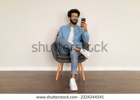 Great App. Young Indian Man Using Smartphone While Sitting In Chair Against White Wall Background, Happy Eastern Male Browsing New Mobile Application Or Messaging Online, Scrolling Social Networks