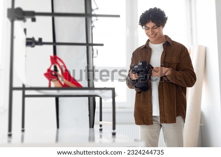 Happy professional photographer man checking the pictures on modern photocamera while doing a content photoshoot, standing near red stylish shoes