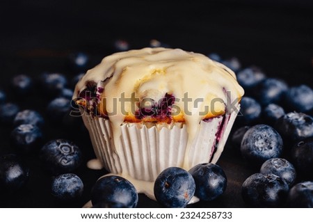Close-up Side View of Blueberry Muffin With Lemon Glaze: Glazed blueberry muffin in a paper wrapper surrounded by fresh blueberries