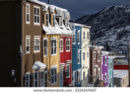 A very steep street in St. John's, Newfoundland, on a foggy winter's day, with bright colored row houses descending down the road. There's snow on the hillside in the background and a smooth harbor. Royalty-Free Stock Photo #2324269607
