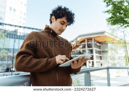 Serious confident latin man holding digital tablet, check email, watching video standing on urban street. Student studying, reading e book in university campus, education concept 