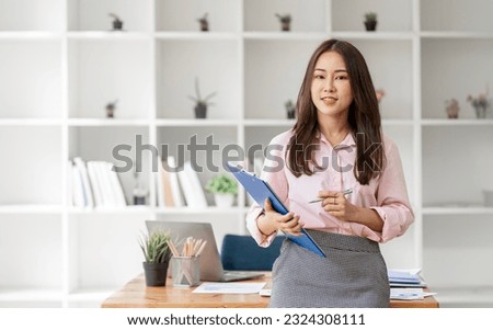 Attractive businesswoman holding file folder standing at office, smiling to camera.