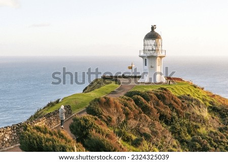 Cape Reinga Lighthouse in New Zealand at sunrise with a woman walking toward it, semi wide angle shot Royalty-Free Stock Photo #2324325039