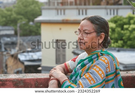 Photo of a simple looking aged Bengali woman (Indian ethnicity) with saddened, depressed look (feeling of loneliness) in her face. Photo taken on a building rooftop, in Kolkata, West Bengal. Royalty-Free Stock Photo #2324352269