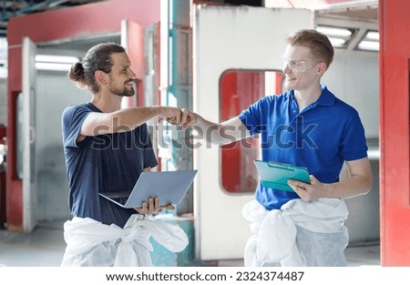Portrait of a professional craftsman Standing with a laptop and a notebook next to a car in front of a car paint shop. Fist-clenched hands when work is done, success with teamwork business