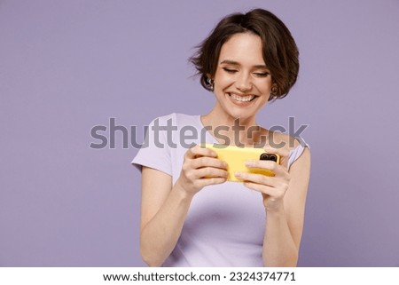Young smiling gambling happy woman 20s wear white t-shirt using play racing app on mobile cell phone hold gadget smartphone for pc video games isolated on pastel purple background studio portrait.