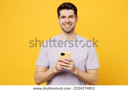 Young smiling happy fun caucasian man he wear light purple t-shirt casual clothes hold in hand use mobile cell phone chat online isolated on plain yellow background studio portrait. Lifestyle concept