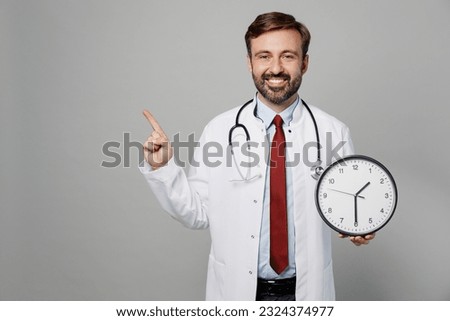 Male doctor man wears white medical gown suit work in hospital hold in hand clock point finger aside on workspace isolated on plain grey color background studio portrait. Healthcare medicine concept