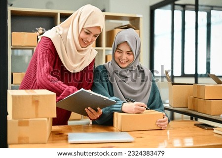 A happy young Asian Muslim female online seller working with her employee in the office. SME business, online shop owner