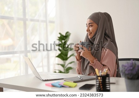 Young charming Muslim businesswoman having a coffee while working with laptop in the office room. 