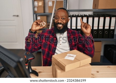 African american man working at small business ecommerce holding bitcoin surprised with an idea or question pointing finger with happy face, number one 