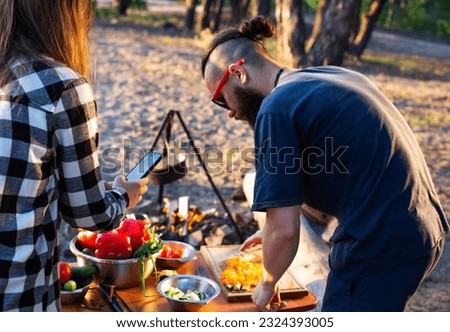 Stylish man with a beard and black glasses cuts fresh vegetables peppers and tomatoes to prepare a salad in nature. Vacation with friends in nature. Tourism as a hobby. Evening sunset, recreation