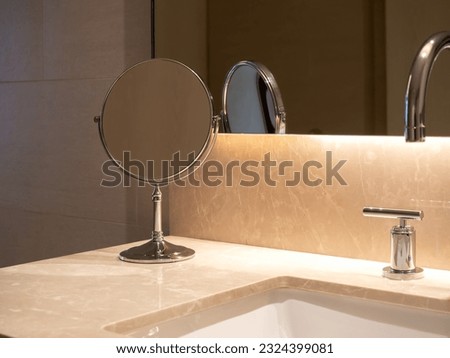 Small folding round makeup mirror standing near sink basin with marble top and steel faucet near the large wall glass window with hidden lighting. Modern design of white clean bathroom interior.
