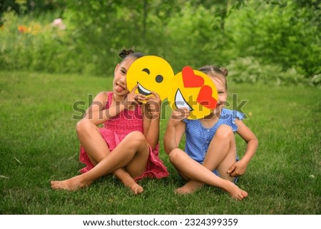 Playful laughing funny girls hide behind a variety of smile faces sitting in the park on a lawn on a sunny summer day
