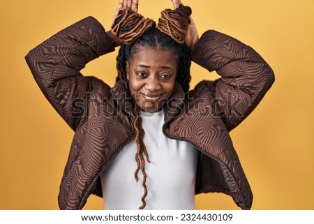 African woman with braided hair standing over yellow background doing bunny ears gesture with hands palms looking cynical and skeptical. easter rabbit concept. 