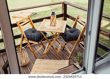 Modern boho style small wood balcony seating corner with wooden garden furniture, soft black pillows, lantern with burning candle on table outdoors in summer. Royalty-Free Stock Photo #2324435711